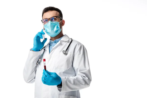 Médecin en masque médical et manteau blanc parlant sur smartphone tout en tenant tube à essai avec échantillon de sang isolé sur blanc — Photo de stock