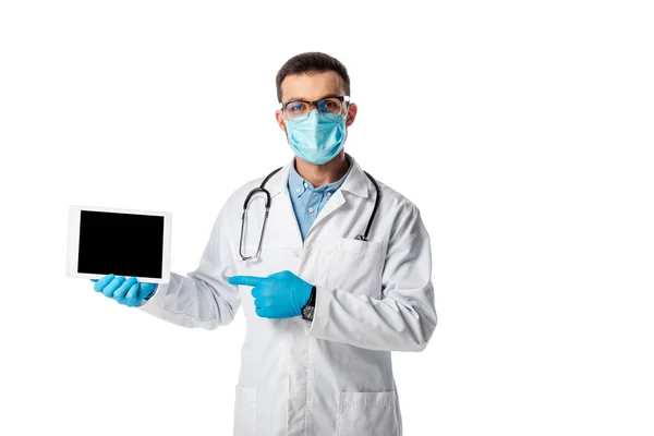 Doctor en máscara médica y bata blanca apuntando con el dedo a la tableta digital con pantalla en blanco aislado en blanco - foto de stock