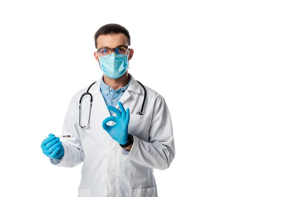 Doctor in medical mask and white coat holding digital thermometer and showing ok sign isolated on white — Stock Photo
