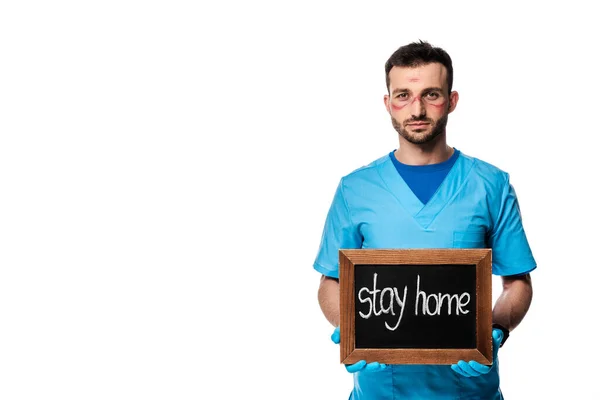 Médico con marcas en la cara que sostiene la pizarra con letras de la estancia en casa aislado en blanco - foto de stock