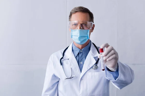 Foyer sélectif du médecin mature dans le masque médical et googles tenant tube à essai avec échantillon de sang sur gris — Photo de stock