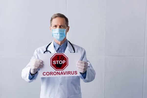 Médico de mediana edad en bata blanca y máscara médica con pancarta con letras de stop coronavirus en gris - foto de stock