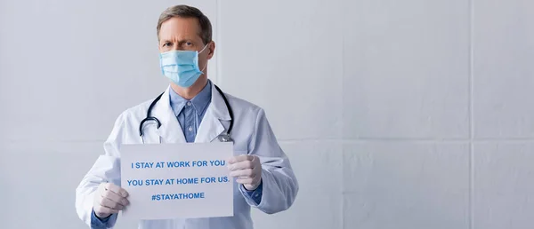 Foto panorámica de médico de mediana edad en máscara médica sosteniendo cartel con me quedo en el trabajo para usted, usted se queda en casa para nosotros las letras en gris - foto de stock