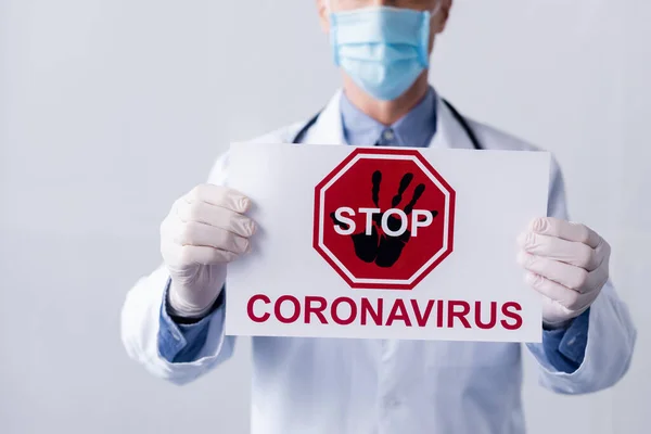 Vista recortada del médico maduro en bata blanca y máscara médica sosteniendo pancarta con letras de coronavirus stop en gris - foto de stock
