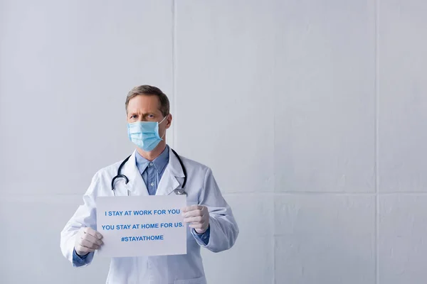Middle aged doctor in medical mask holding placard with i stay at work for you, you stay at home for us lettering on grey — Stock Photo