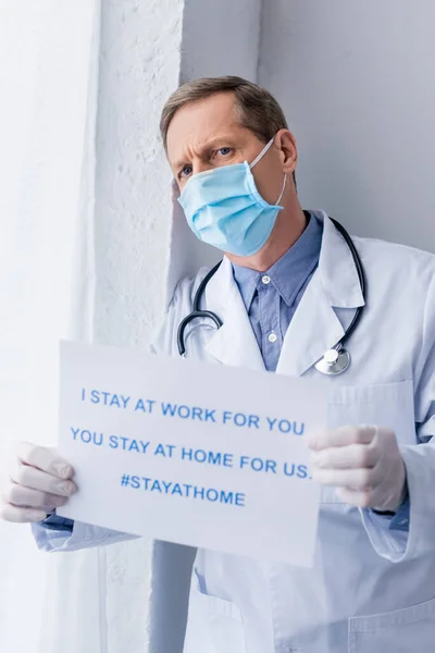 Médico de mediana edad en guantes de látex con pancarta con me quedo en el trabajo para usted, usted se queda en casa para nosotros las letras - foto de stock