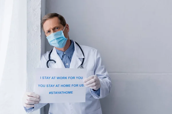 Middle aged doctor in medical mask and latex gloves holding placard with i stay at work for you, you stay at home for us lettering on grey — Stock Photo