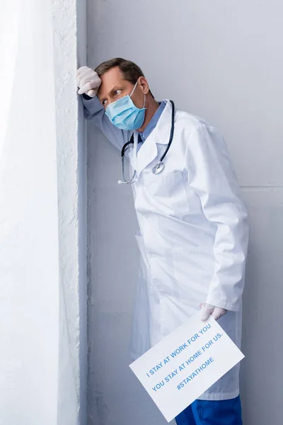 Tired middle aged doctor in medical mask holding placard with i stay at work for you, you stay at home for us lettering on grey — Stock Photo