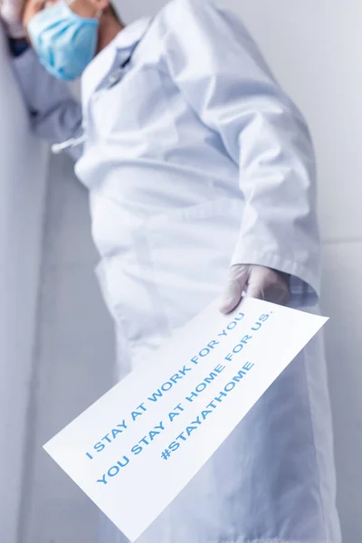 Baixo ângulo vista de cansaço médico de meia-idade em máscara médica segurando cartaz com eu ficar no trabalho para você, você ficar em casa para nós lettering em branco — Fotografia de Stock