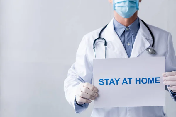 Vista recortada del médico de mediana edad en guantes de látex que sostienen pancarta con letras de estancia en casa en gris - foto de stock