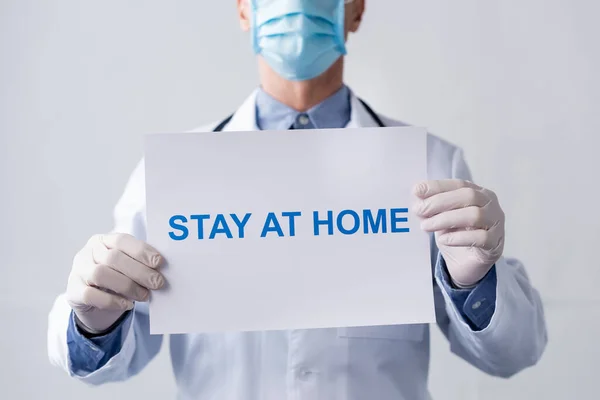 Vista recortada del médico de mediana edad en la máscara médica que sostiene la pancarta con la estancia en casa con letras en gris - foto de stock