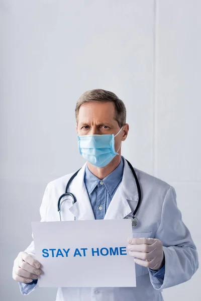 Médico de mediana edad en máscara médica sosteniendo cartel con la estancia en casa con letras en gris - foto de stock
