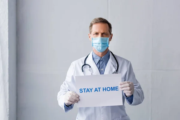 Mature doctor in medical mask and latex gloves holding placard with stay at home lettering on grey — Stock Photo