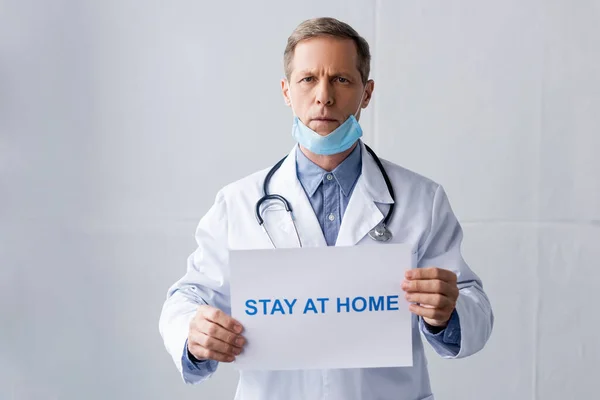 Medico maturo in maschera medica tenuta cartello con soggiorno a casa lettering su grigio — Foto stock