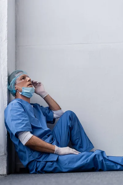 Tired and mature doctor in medical cap and goggles sitting near white wall — Stock Photo
