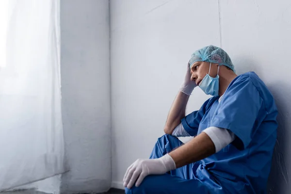 Médico cansado y maduro en gorra médica y gafas sentado cerca de la pared blanca en la clínica - foto de stock