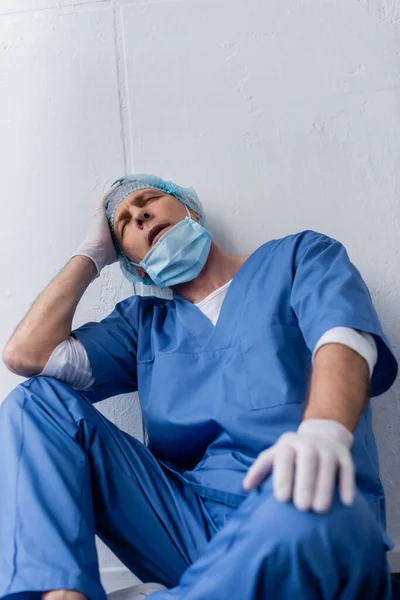 Tired and mature doctor with closed eyes sitting near white wall — Stock Photo