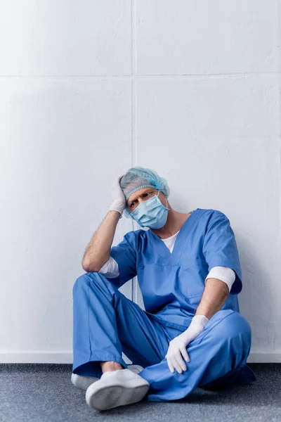 Tired and mature doctor in medical mask and cap sitting on white — Stock Photo