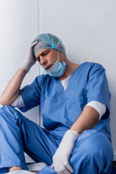 Selective focus of exhausted and mature doctor in medical mask and cap sitting on white — Stock Photo