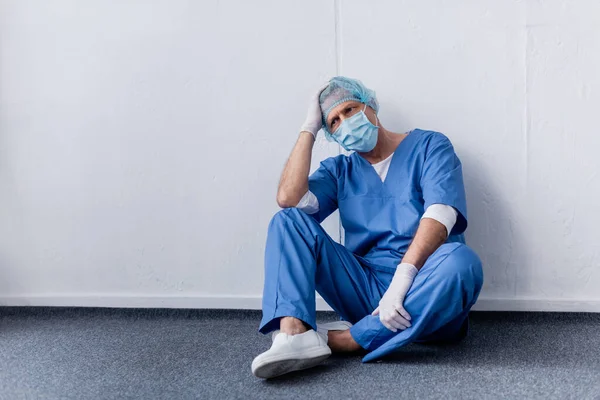 Médico cansado y de mediana edad en gorra médica y gafas sentado cerca de la pared blanca - foto de stock