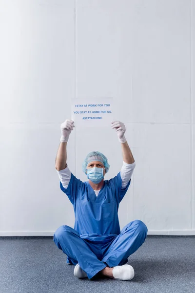 Médico de mediana edad en máscara médica y la tapa de la celebración de la pancarta con me quedo en el trabajo para usted, usted se queda en casa para nosotros las letras mientras está sentado en blanco - foto de stock