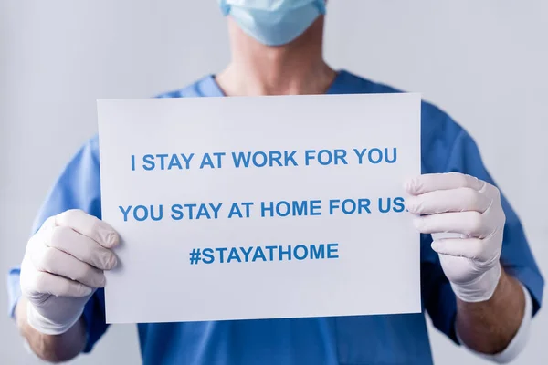 Cropped view of mature doctor holding placard with i stay at work for you, you stay at home for us lettering on white — Stock Photo