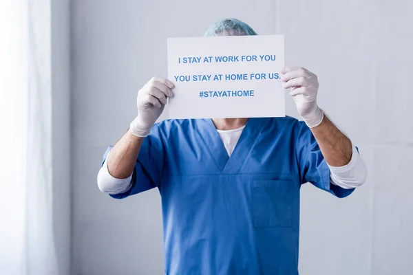 Mature doctor covering face with placard with i stay at work for you, you stay at home for us lettering on white — Stock Photo