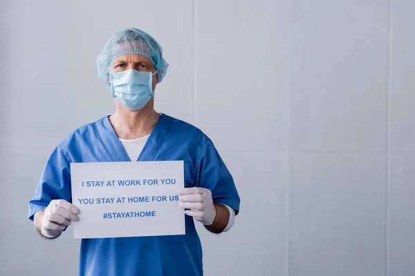 Médico maduro en máscara médica y el tablero de la tapa con me quedo en el trabajo para usted, usted se queda en casa para nosotros las letras en gris - foto de stock