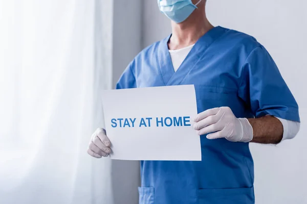 Обрізаний вид лікаря середнього віку в медичній масці, що тримає плакат з перебуванням вдома — стокове фото