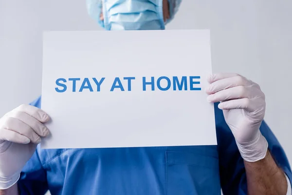 Vista ritagliata del medico di mezza età che tiene cartello con soggiorno a casa lettering sul grigio — Foto stock