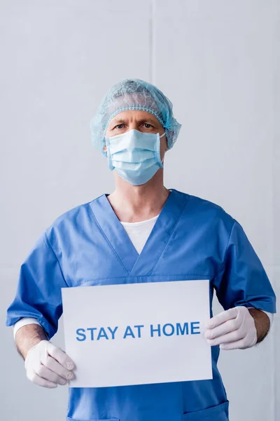 Medico maturo in maschera medica e tappo con cartello con soggiorno a casa lettering su grigio — Foto stock