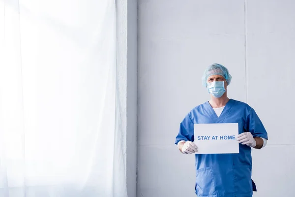 Médico maduro en máscara médica, gorra y guantes de látex sosteniendo pancarta con letras de estancia en casa - foto de stock