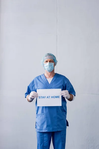 Médico maduro em máscara médica, tampa e luvas de látex segurando cartaz com estadia em casa lettering e em pé sobre cinza — Fotografia de Stock