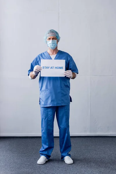 Médico maduro en máscara médica, gorra y guantes de látex sosteniendo pancarta con letras de estancia en casa mientras está de pie en gris - foto de stock