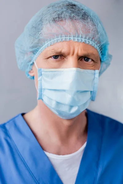 Mature doctor in blue medical mask and cap looking at camera on grey — Stock Photo