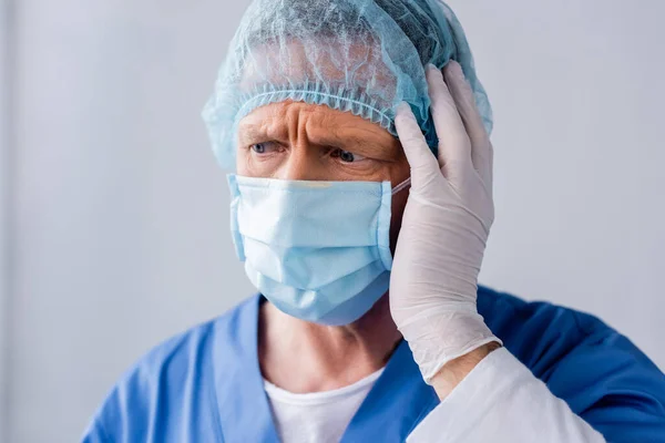 Tired and mature doctor in blue medical mask touching medical cap on grey — Stock Photo