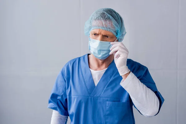 Зрілий лікар торкається синьої медичної маски на сірому — стокове фото