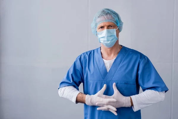 Зрілий лікар у блакитній медичній масці, кепці та латексних рукавичках зі згорнутими руками на сірому — стокове фото