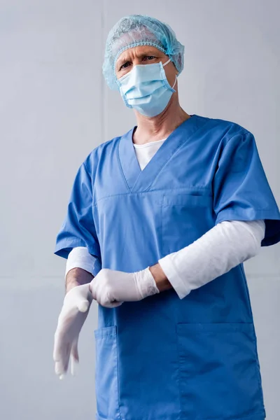 Mature doctor in blue medical mask and cap wearing latex glove on grey — Stock Photo