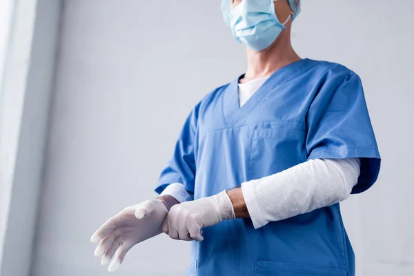 Cropped view of mature doctor in blue medical mask wearing latex glove on grey — Stock Photo