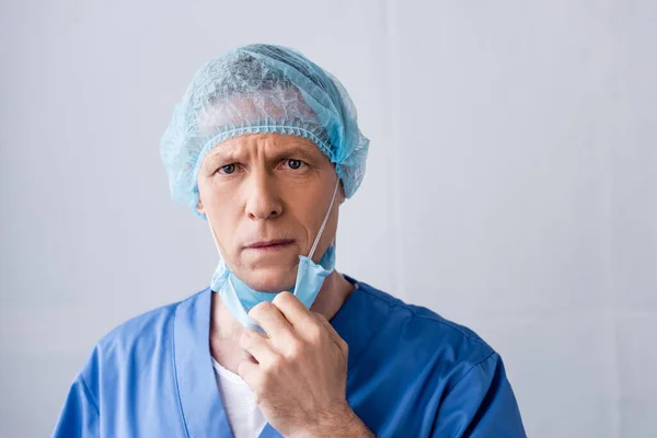 Médico maduro en azul casquillo médico tocando máscara médica en gris - foto de stock