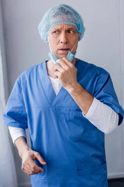 Médico maduro en la tapa médica azul tocando máscara médica y mirando hacia otro lado - foto de stock