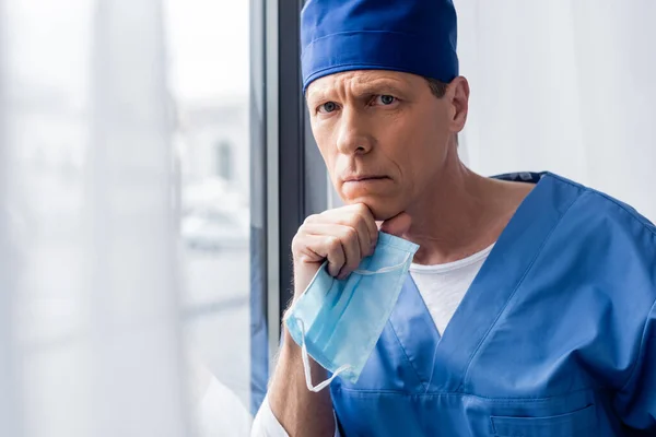 Médico pensativo e maduro em chapéu esfoliante azul segurando máscara médica e olhando para a câmera — Fotografia de Stock