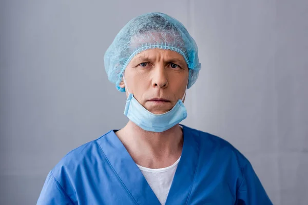 Зрілий лікар у блакитній медичній шапці та масці дивиться на камеру на сірому — стокове фото