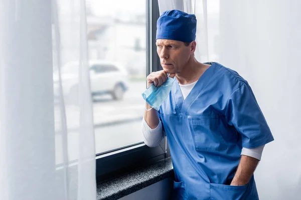 Medico pensieroso e maturo in camice blu con maschera medica, in piedi con la mano in tasca e guardando la finestra — Foto stock