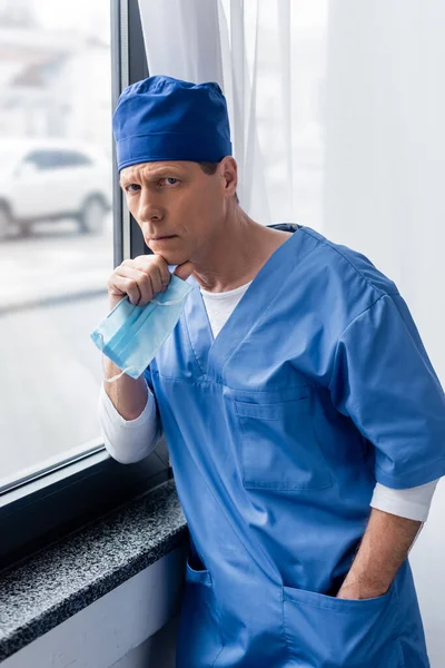 Médico maduro en sombrero de matorral azul que sostiene la máscara médica, de pie con la mano en el bolsillo y mirando a la cámara cerca de la ventana - foto de stock
