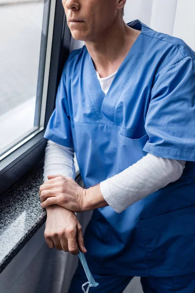 Ausgeschnittene Ansicht eines Arztes mittleren Alters mit medizinischer Maske, der am Fenster im Krankenhaus steht — Stockfoto