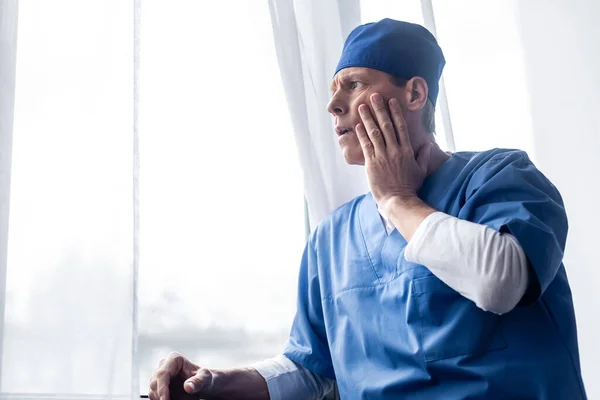 Tired and middle aged doctor in scrub hat standing near window in hospital and touching face — Stock Photo