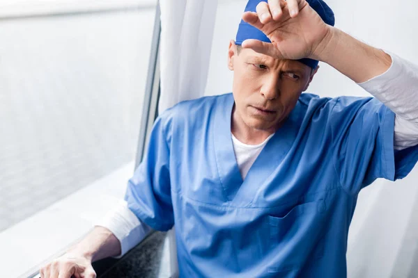 Médecin fatigué et d'âge moyen dans le chapeau de gommage touchant le front près de la fenêtre à l'hôpital — Photo de stock