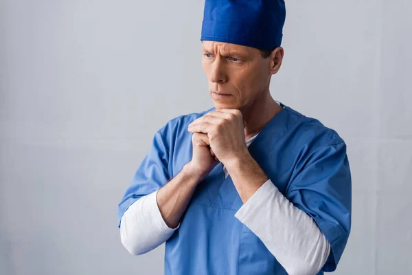 Middle aged doctor in scrub hat standing with clenched hands while thinking on grey — Stock Photo